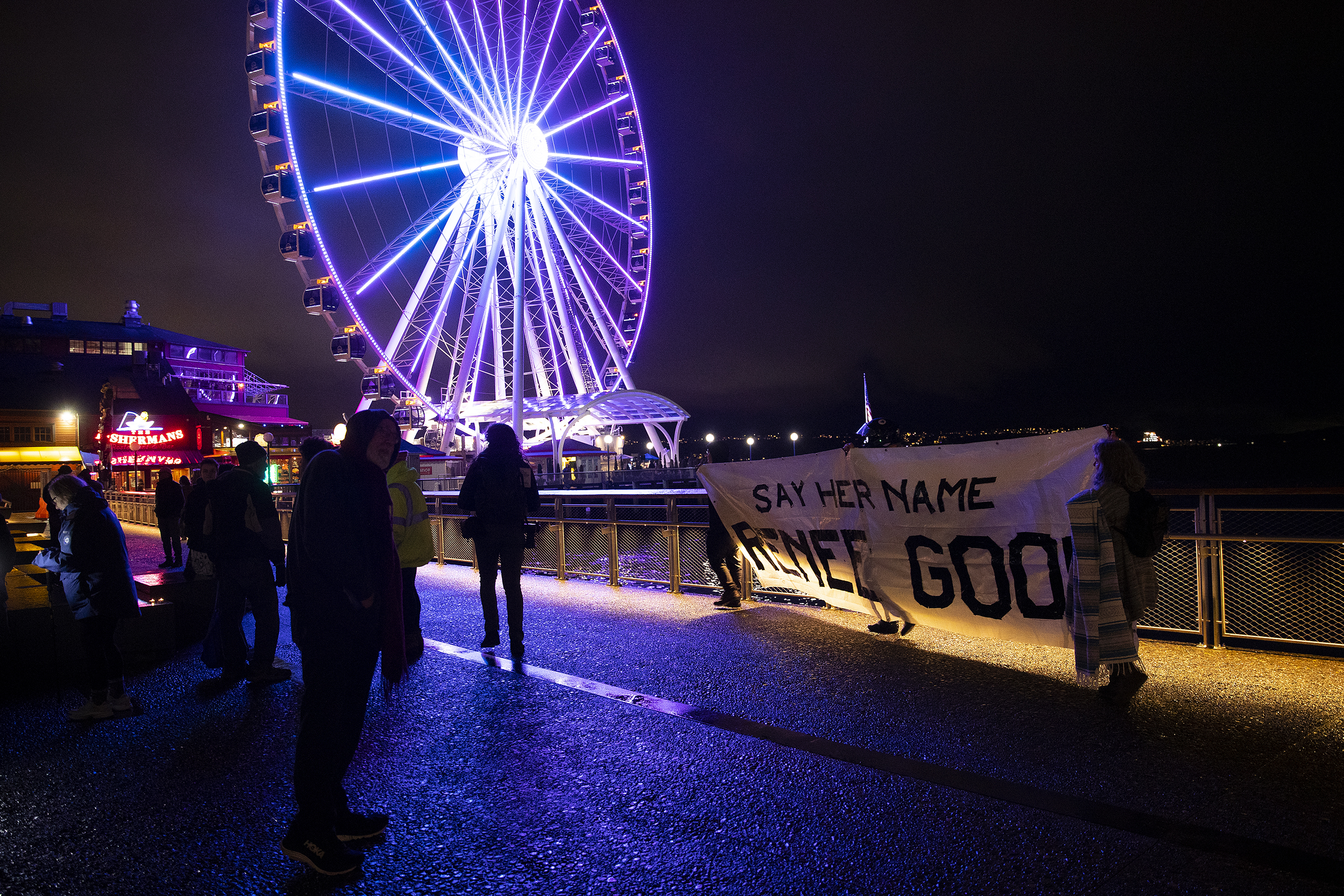 caption: A banner reads ‘Say Her Name, Renee Good’ as hundreds gather to honor the Minneapolis mother of three who was shot in the head and killed by an ICE agent earlier this week, on Thursday, January 8, 2026, at Pier 58 in Seattle. 