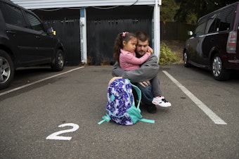caption: Sean holds Vay, 5, as they wait for the school bus on the first day of school on Monday, September 12, 2022, outside of their apartment complex in Lynnwood.