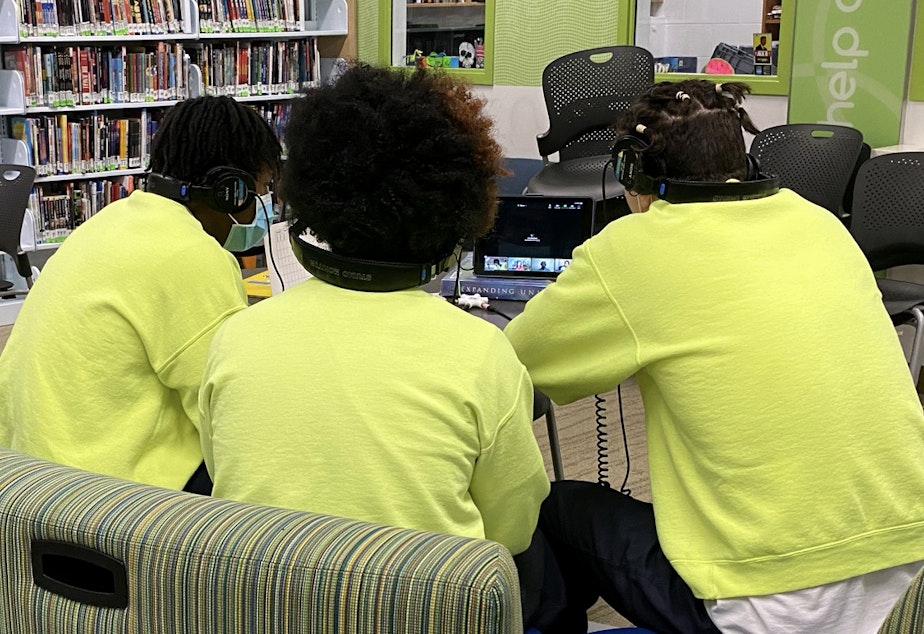 caption: Three teens wearing headphones huddle close together to see their shared iPad during a virtual RadioActive workshop at the Judge Patricia H. Clark Children and Family Justice Center on April 14, 2021.