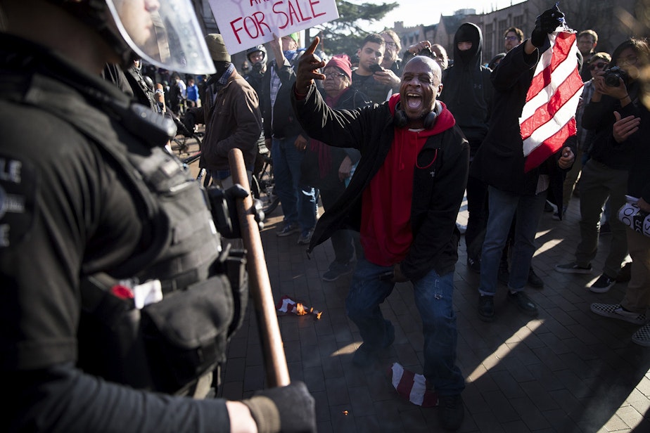 caption: Jamal X, center, yells at police officers after lighting a portion of an American Flag on fire while protesting outside of a College Republicans rally on Saturday, Feb. 10, 2018, at Red Square on the University of Washington campus in Seattle.