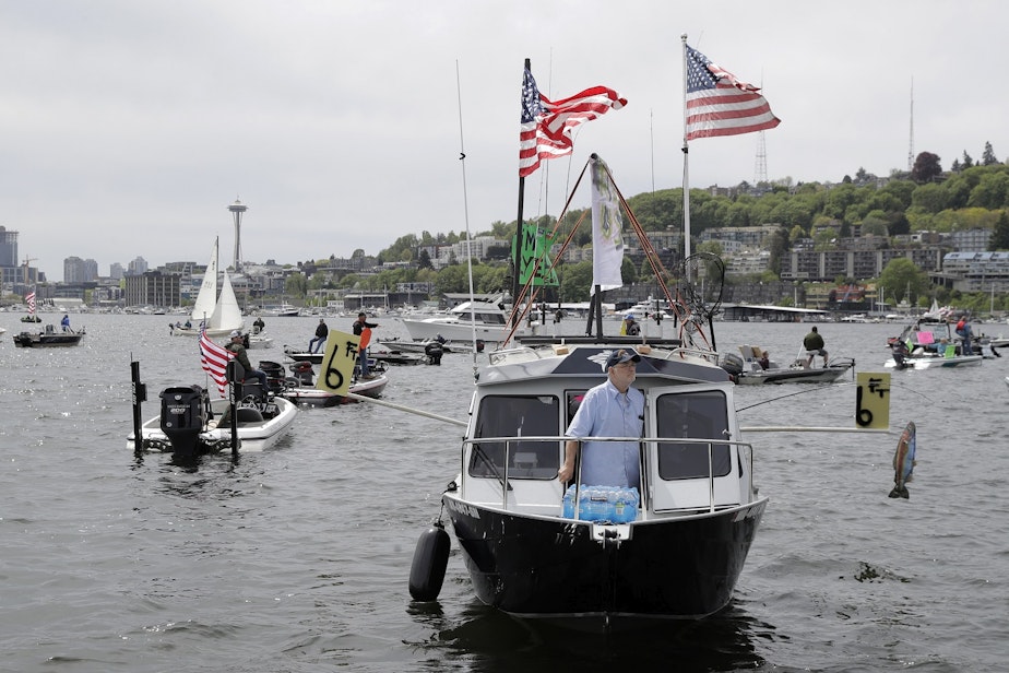 caption: A protester reels in a fake fish near poles marking a six-foot social distance on a boat on Lake Union near Gas Works Park in Seattle, Sunday, April 26, 2020, during a protest against Washington state's current ban on fishing due to stay-at-home orders implemented to prevent the spread of the coronavirus.
