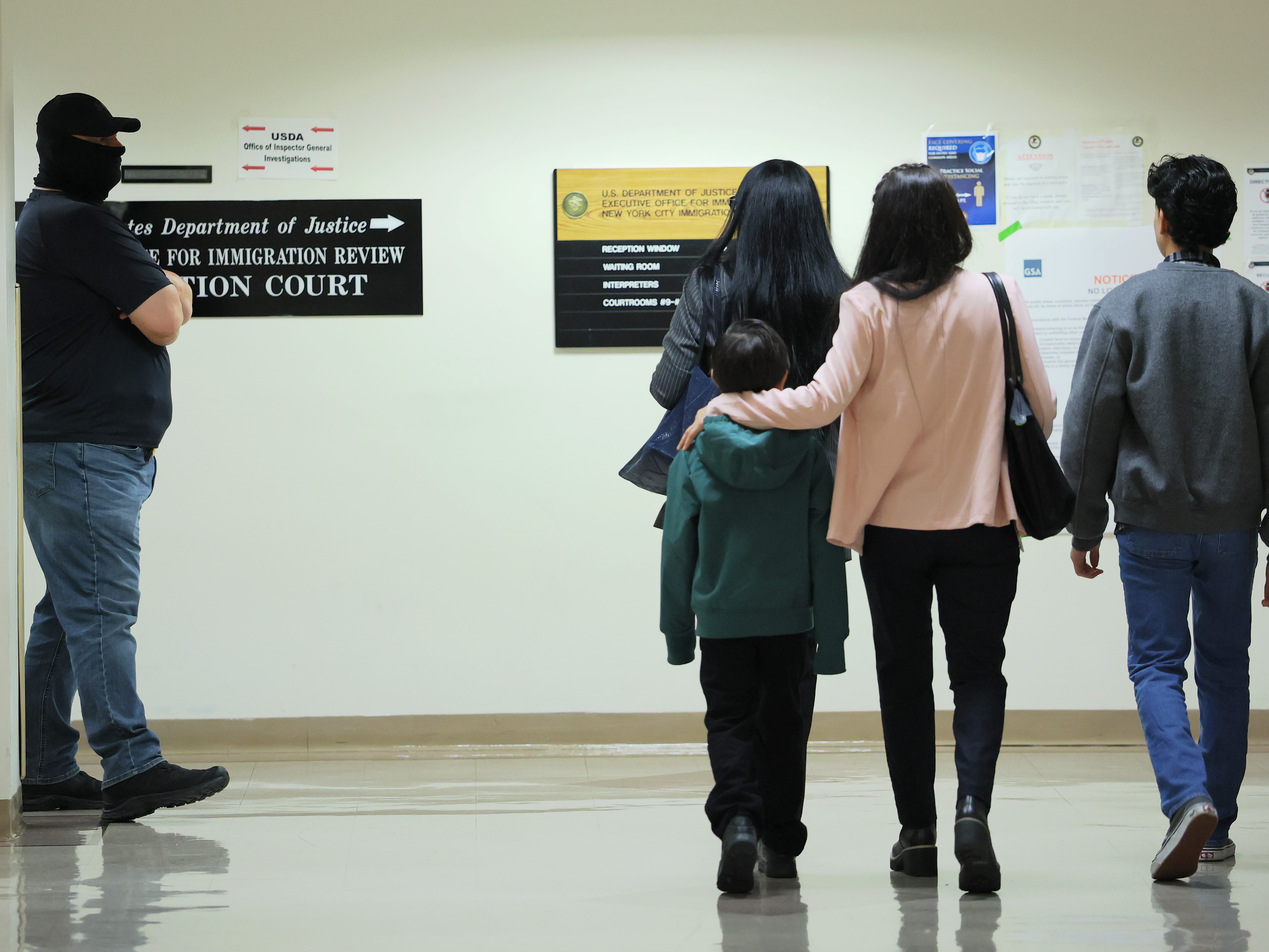caption: People walk past a federal agent as he patrol the halls of immigration court at the Jacob K. Javits Federal Building on Oct. 15 in New York City. Michael M. Santiago/Getty Images