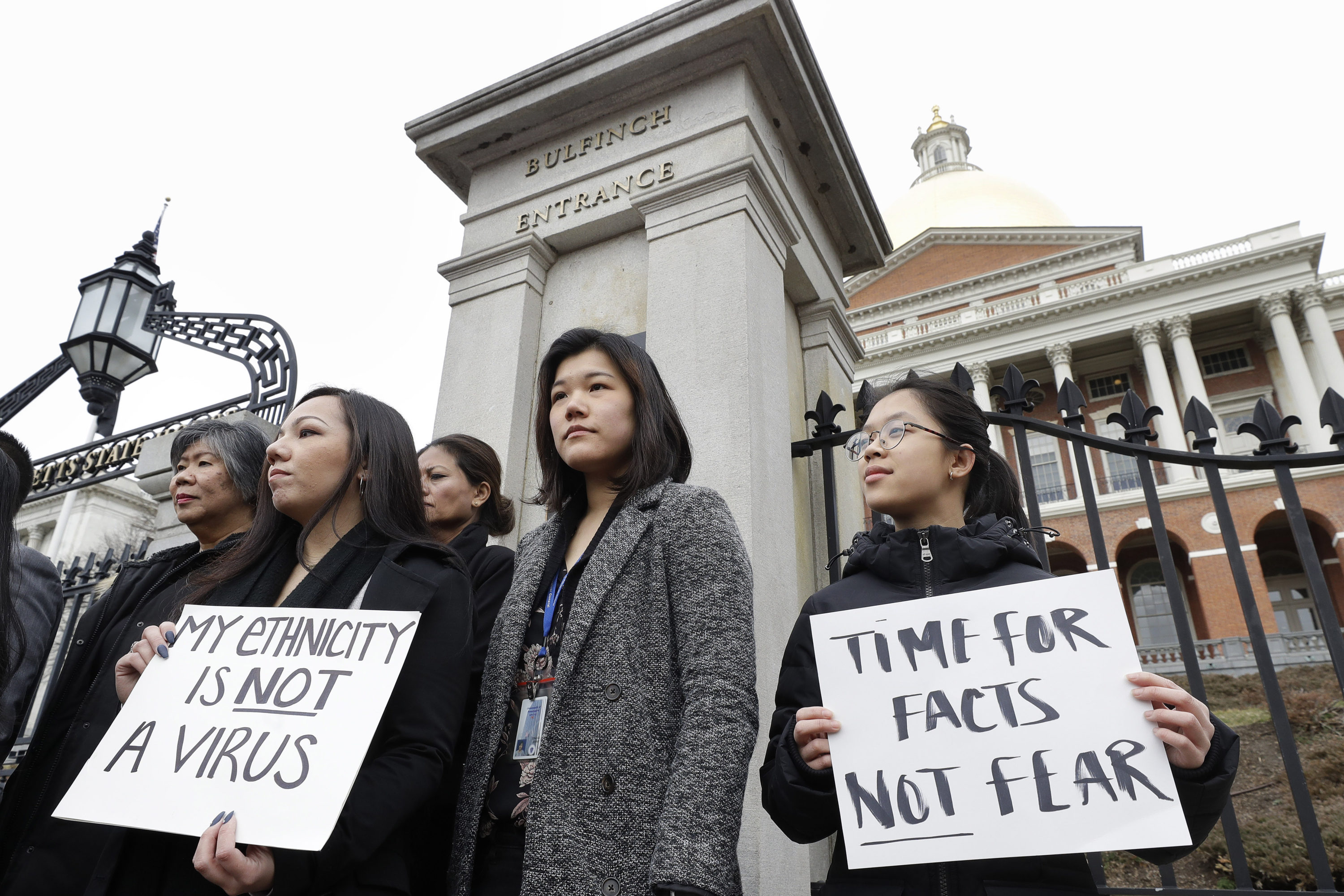 caption: Jessica Wong, of Fall River, Mass., front left, Jenny Chiang, of Medford, Mass., center, and Sheila Vo, of Boston, from the state's Asian American Commission, stand together during a protest, Thursday, March 12, 2020, on the steps of the Statehouse in Boston. (Steven Senne/AP)