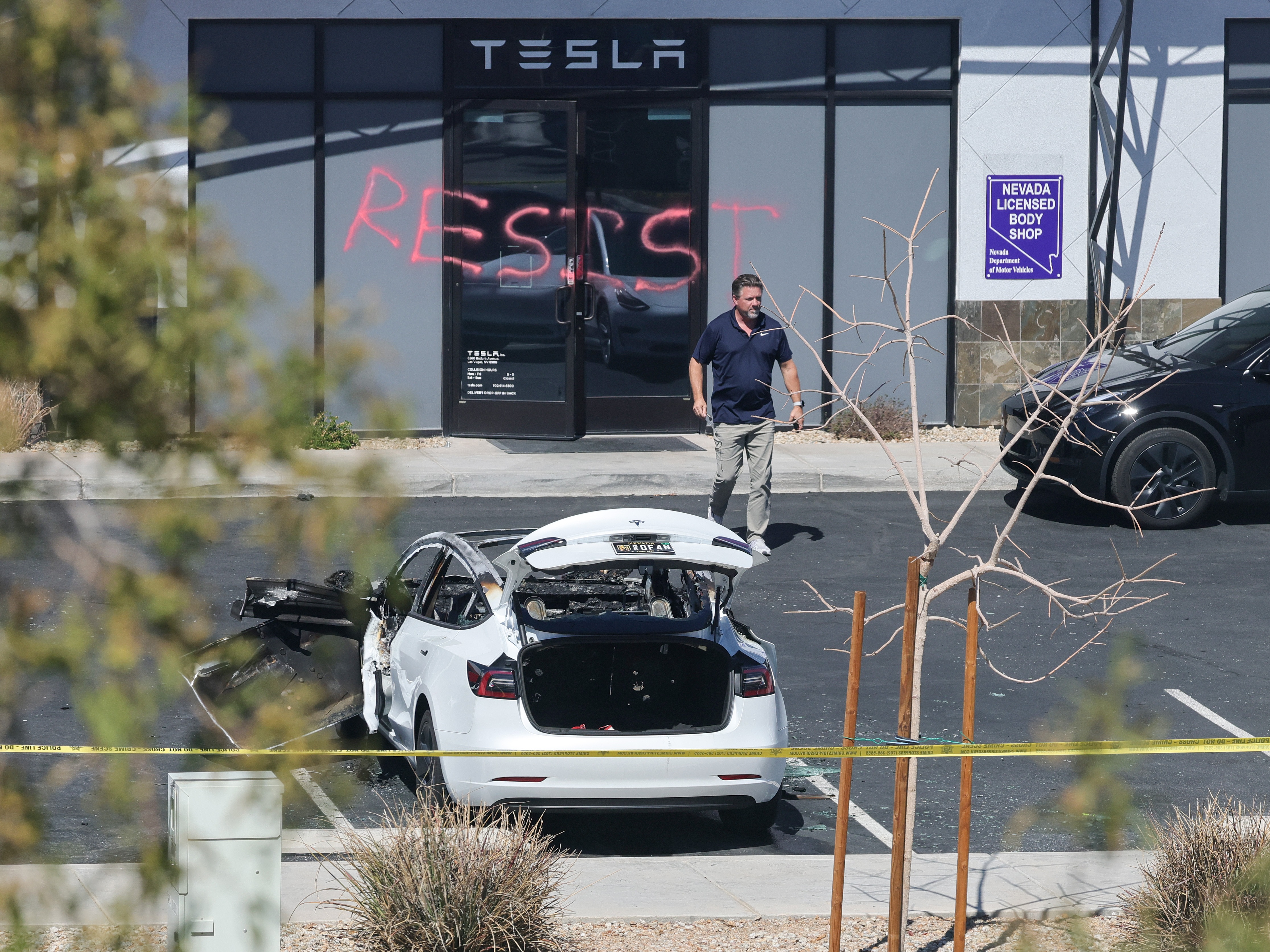 caption: An investigator looks over the scene at a Tesla Collision Center after a person used incendiary devices to set several vehicles on fire on Tuesday in Las Vegas. The Las Vegas Metropolitan Police Department said that five Teslas were damaged and believe that the suspect fired three rounds from a firearm at the vehicles and spray painted the word "RESIST" on the entrance.