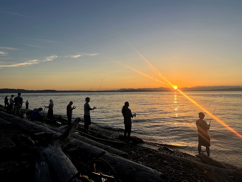 caption: Several people fish along the shore of Lincoln Park as the sun sets on Wednesday, August 16, 2023, in Seattle. 