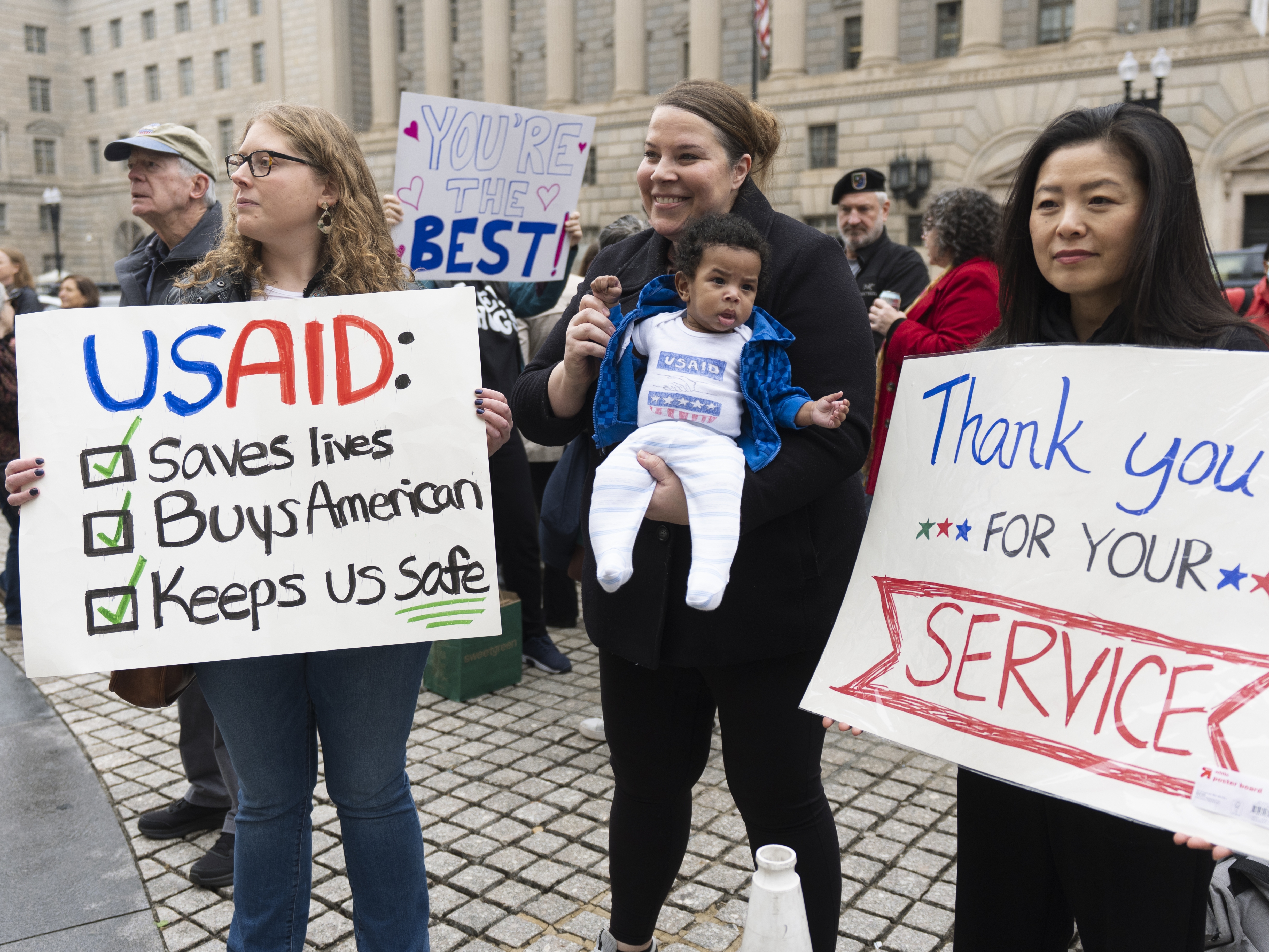 caption: United States Agency for International Development (USAID) supporters hold banners as USAID workers retrieve their personal belongings from the USAID's headquarters in Washington, Thursday, Feb. 27, 2025.