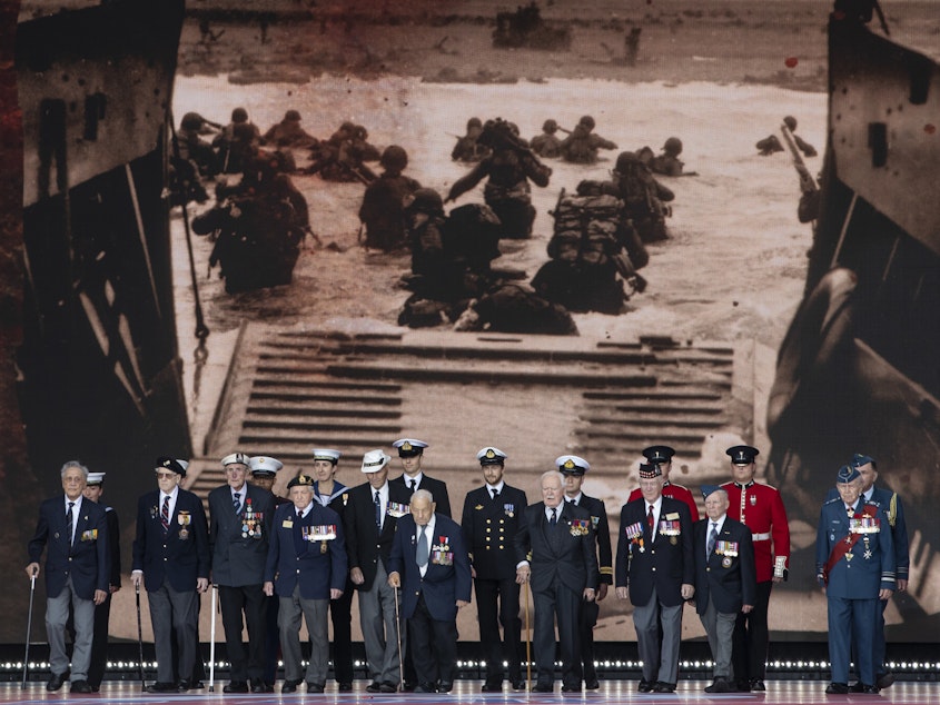 caption: Veterans stand in front of a 1944 image of their comrades wading onto the beaches of France during D-Day commemorations in Portsmouth, England. Leaders of 16 countries involved in World War II joined Queen Elizabeth II at the ceremony on Wednesday.