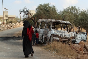 caption: A woman walks past a wrecked van near the northwestern Syrian village of Barisha. Local residents and medical staff tell NPR that noncombatant civilians were killed and injured in the van the night of the U.S. raid on the compound of ISIS leader Abu Bakr al-Baghdadi.
