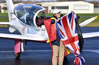 caption: Zara Rutherford, 19, carries the Belgian and British flags on the tarmac after landing her Shark ultralight plane at the Kortrijk airport in Belgium, on Thursday at the completion of a record-breaking solo circumnavigation.