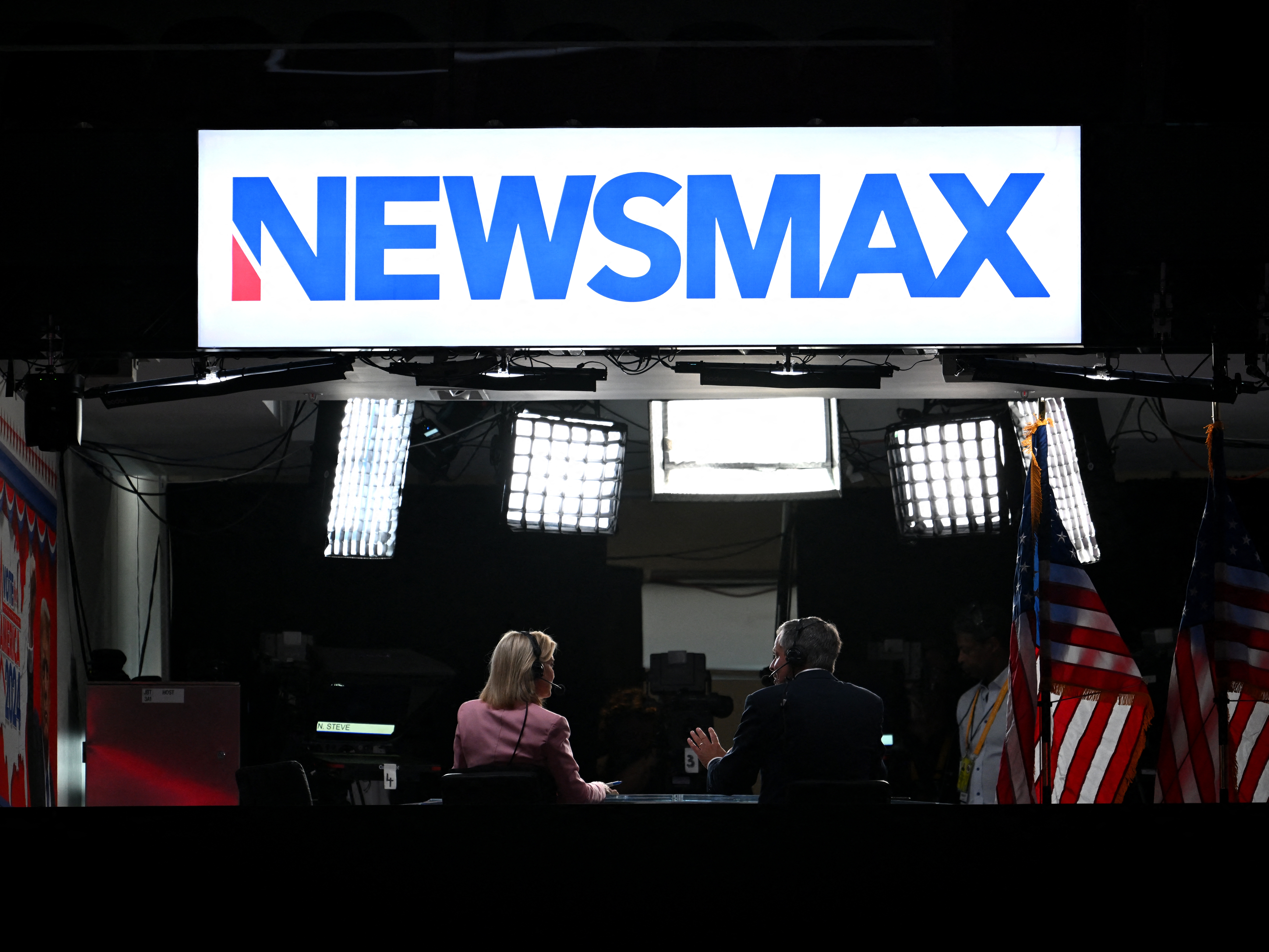 caption: News anchors work at Newsmax's booth during the 2024 Republican National Convention in Milwaukee, Wisconsin. The network has settled with voting-tech company Smartmatic, which accused it of defamation following the 2020 presidential election. 