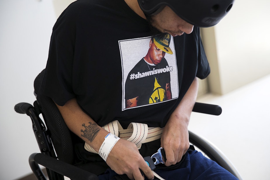 caption: DaShawn Horne buckles the belt of his wheelchair while wearing a t-shirt with his picture and the words #shawniswoke, on Thursday, April 19, 2018, during physical therapy at Harborview Medical Center in Seattle. 