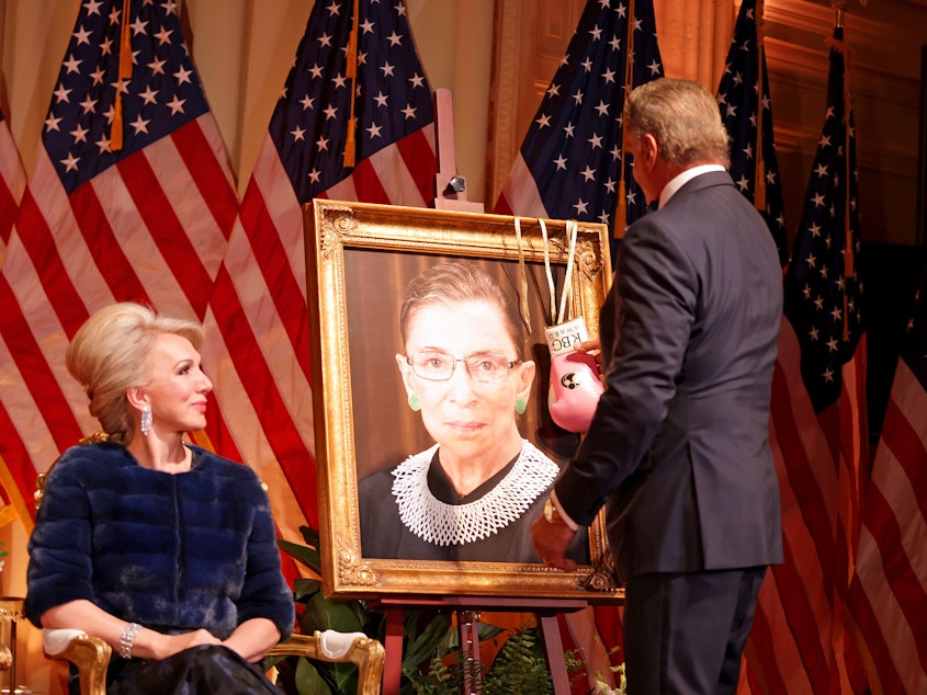 caption: Sylvester Stallone hangs a pair of pink boxing gloves on a portrait of the late Supreme Court Justice Ruth Bader Ginsburg at the Justice Ruth Bader Ginsburg Woman of Leadership Award in 2022, as Julie Opperman, chair of the Dwight D. Opperman Foundation, looks on.