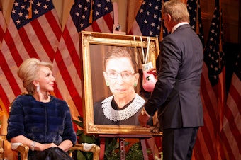 caption: Sylvester Stallone hangs a pair of pink boxing gloves on a portrait of the late Supreme Court Justice Ruth Bader Ginsburg at the Justice Ruth Bader Ginsburg Woman of Leadership Award in 2022, as Julie Opperman, chair of the Dwight D. Opperman Foundation, looks on.