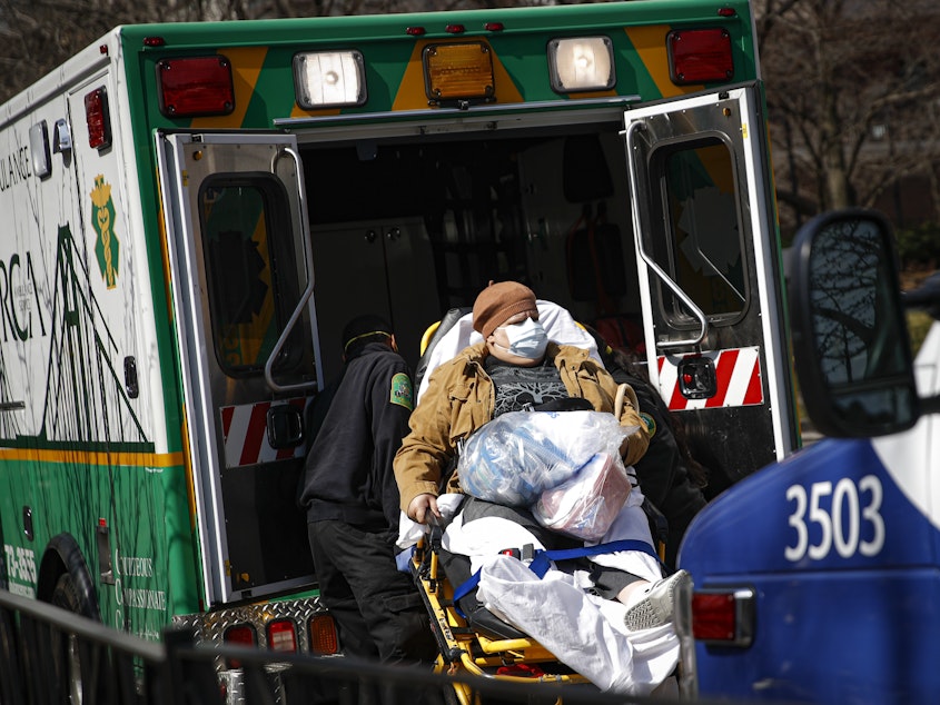 caption: Medical workers load a patient wearing a protective mask into the back of an ambulance Wednesday outside a hospital in Brooklyn. New York has seen a spike in coronavirus cases, and officials are preparing for an influx at medical centers throughout the state.