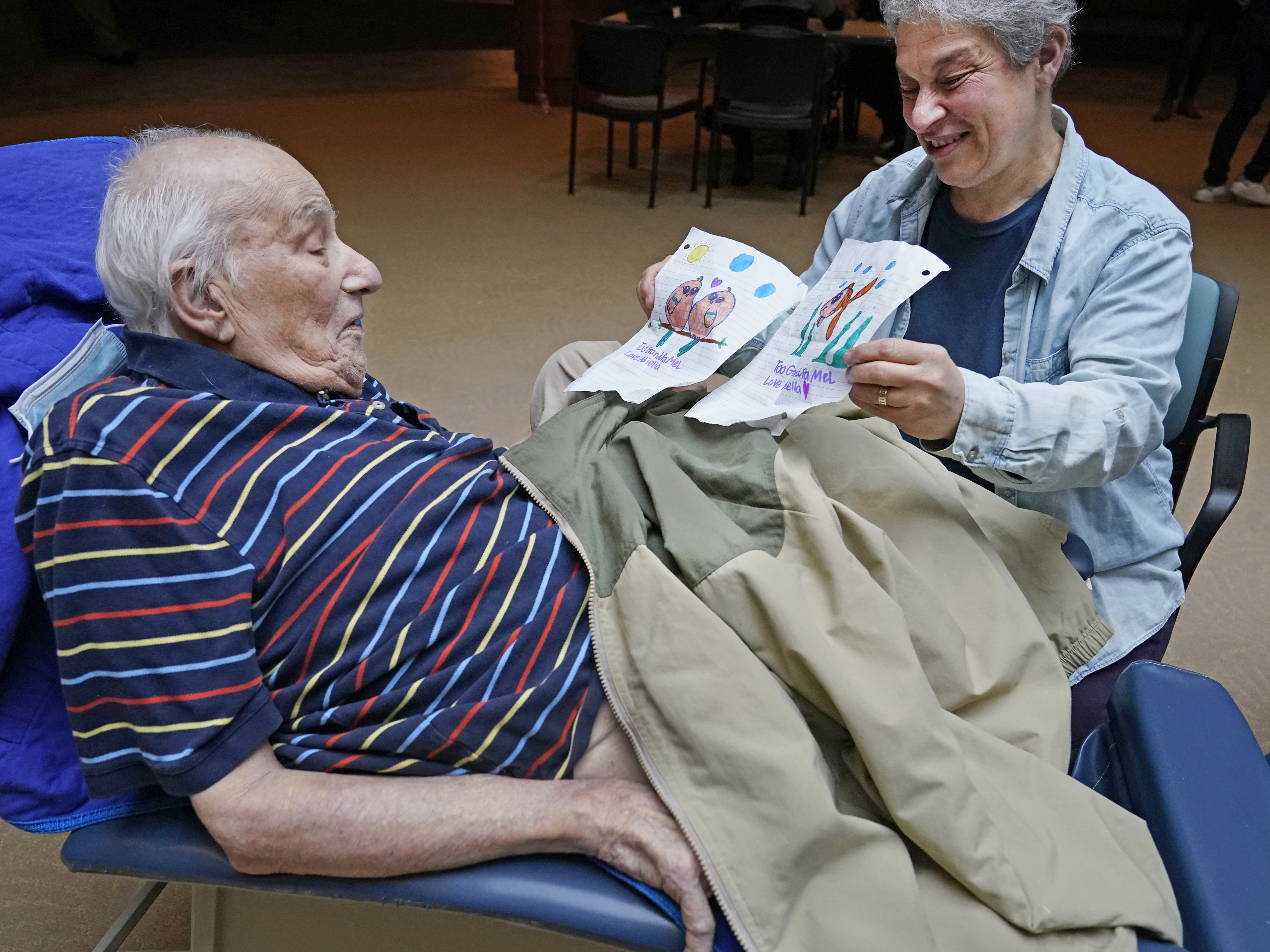 caption: Melvin Goldstein, 90, glances at pictures of birds, left, and a fish, his 13-year-old granddaughter drew for him as a gift as his daughter Barbara Goldstein shares them with his during a family visit inside the Hebrew Home at Riverdale on March 28 in New York. On Friday, the government eased many remaining pandemic restrictions.