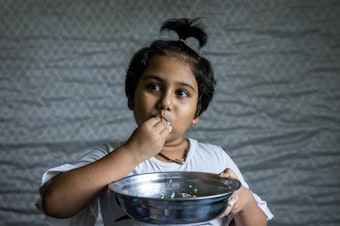 caption: Hosna Ali, 6, eats rice with dried fish at home in Seleyang, Malaysia. Her dad, Mohd Ali, says the family has not been getting enough nutrients during the pandemic.