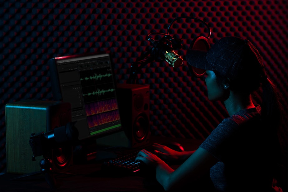 caption: In this stock photo, a young adult wearing a t-shirt sits in a darkened podcast recording studio.