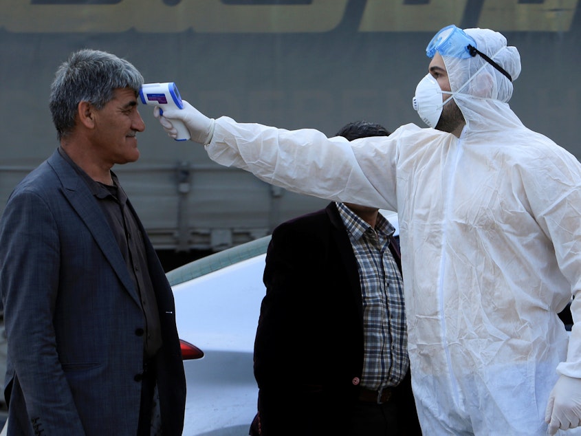 caption: As the coronavirus that causes COVID-19 spreads to more areas, a health worker checks a man's temperature at a checkpoint in the outskirts of Duhok, Iraq, on Tuesday.