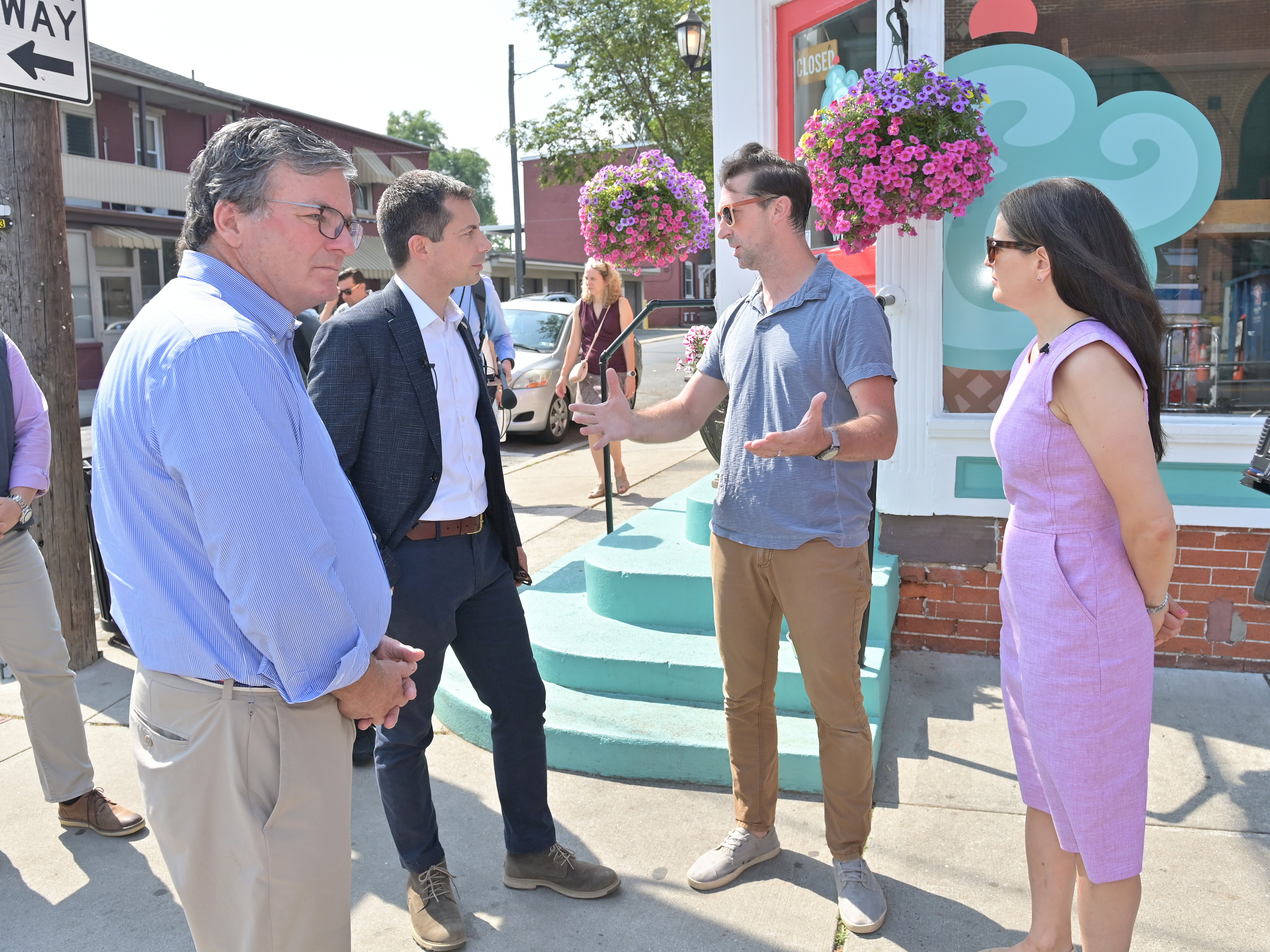 caption: From left, Pennsylvania Secretary of Transportation Mike Carroll, U.S. Secretary of Transportation Pete Buttigieg, Dave Brown, owner of Splits and Giggles Cafe, and Lancaster, Pa., Mayor Danene Sorace speak next to one of the city's new protected bike lanes in June 2024. 