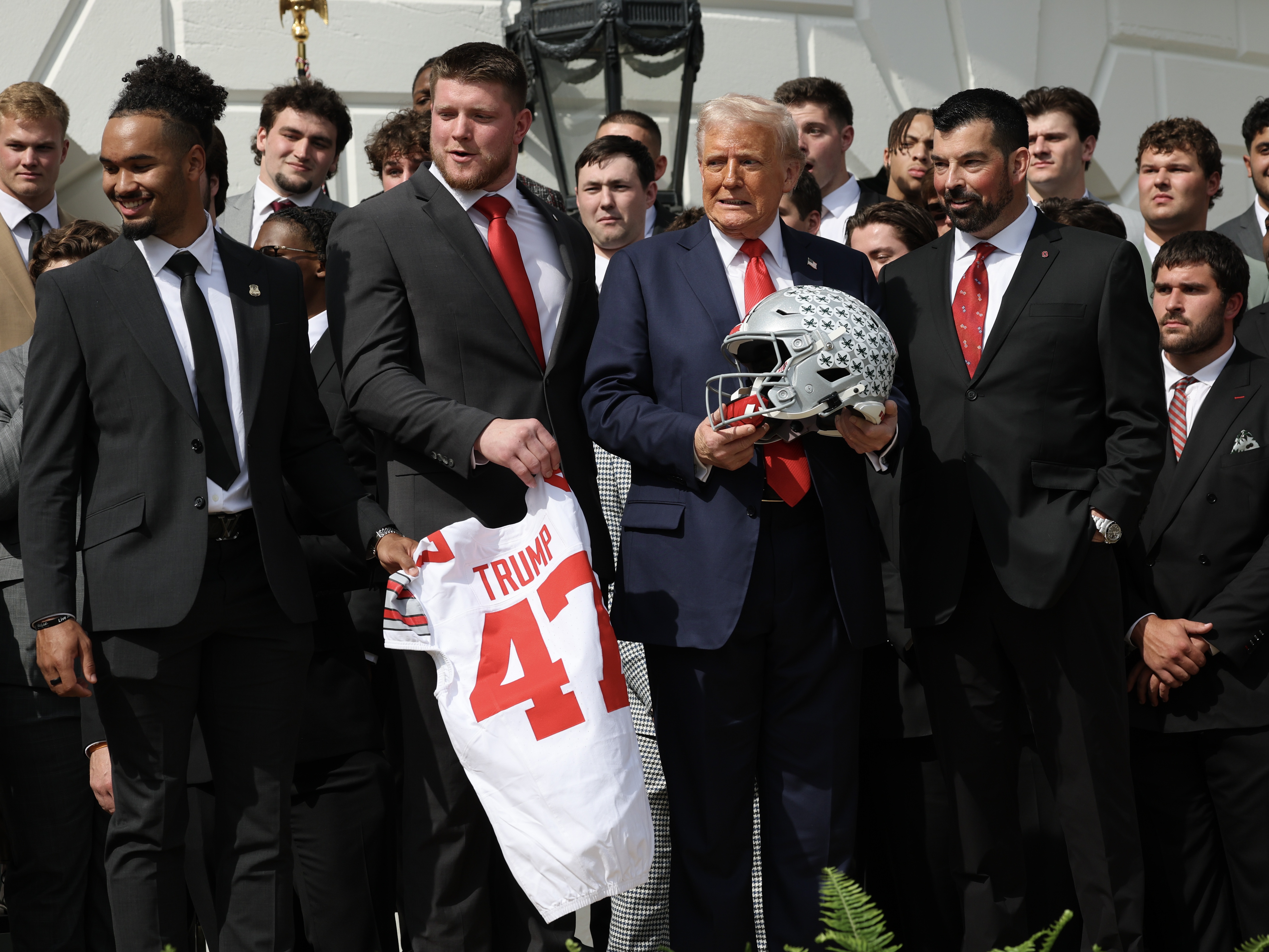 caption: President Trump stands with Ohio State Head Coach Ryan Day (R) as he welcomed the 2025 College Football National Champions from Ohio State University to the White House during a ceremony on April 14, 2025 in Washington, DC. Trump's new executive order seeks to ban "pay-for-play" deals and requires schools to provide scholarships in women's and Olympic sports — and it dangles the threat of withholding federal funds to schools as an enforcement mechanism.