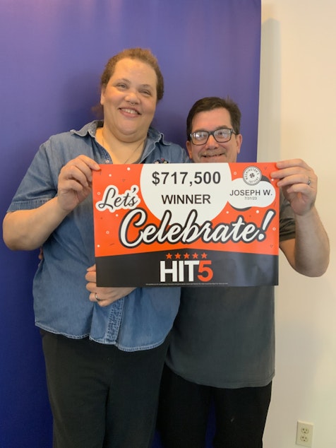 caption: Joseph Waldherr and his wife hold up a promotional sign celebrating their Hit 5 winning ticket.