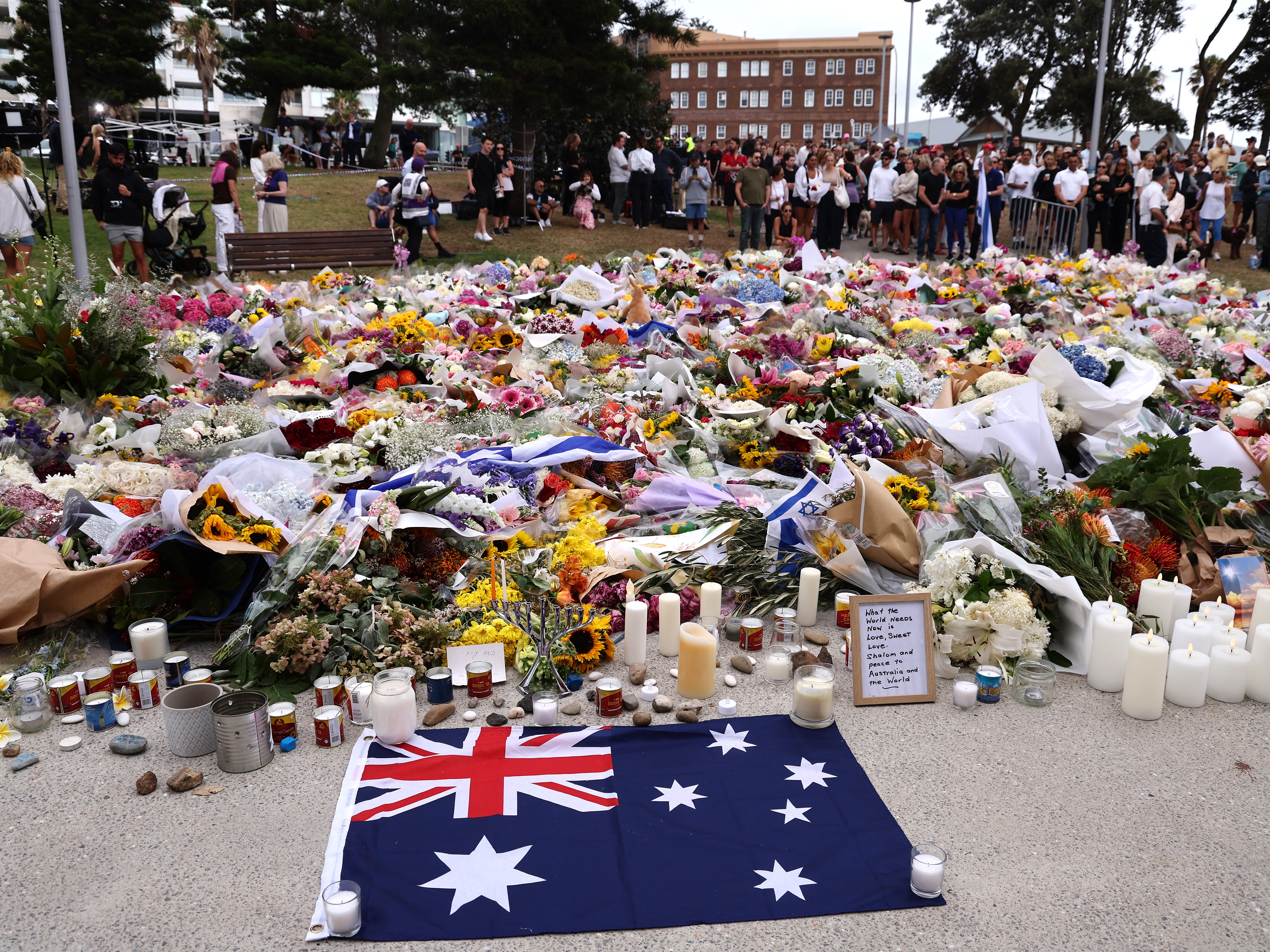 caption: Mourners gather around floral tributes at Bondi Pavilion to honor the victims of the Bondi Beach shooting in Sydney on December 16, 2025. A father-and-son team toting long-barrelled guns shot and killed 15 people including a 10-year-old girl at Sydney's Bondi Beach on December 14, with authorities labelling it an antisemitic terrorist attack on a Jewish festival.