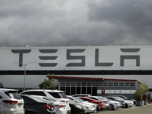 caption: Vehicles are parked outside the Tesla plant in Fremont, Calif., on May 12, 2020. A federal judge has slashed the award to a Black former contract worker over claims that he was subjected to racial discrimination at the factory.