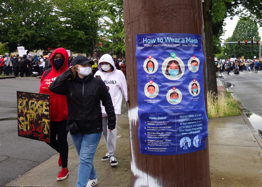 caption: Demonstrators departing the silent march pass a flyer detailing the proper way to wear a mask. Despite concerns that protests would lead to a spike in COVID-19 infections in King County, preliminary testing of protesters suggests that the safety precautions taken by march organizers were successful in limiting the virus' spread among marchers. At the June 12th protest, masks were mandatory.