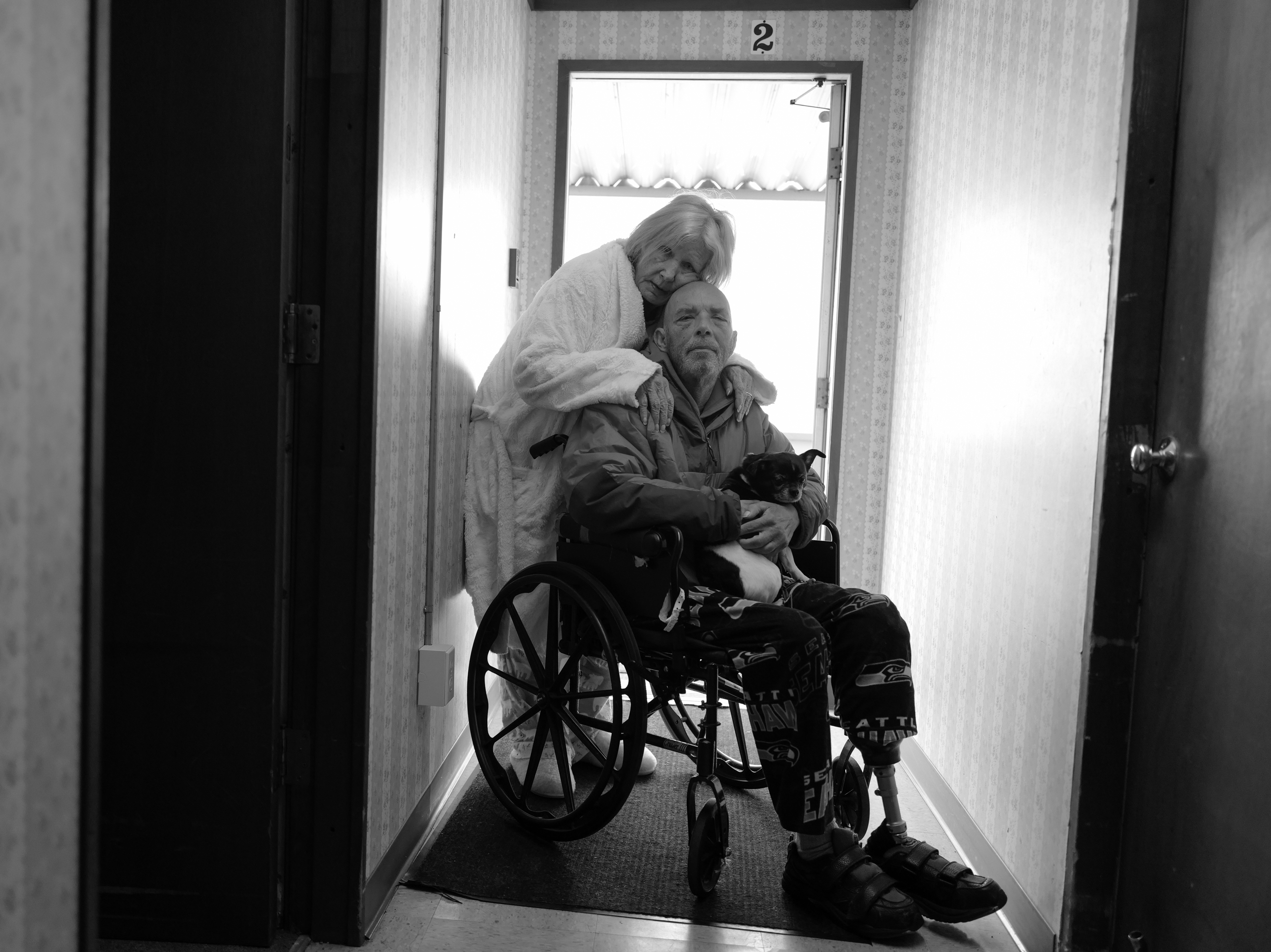 caption: Karen hugs Tony in his wheelchair on April 14, 2022. They now live in Burlington, Wash., and their mobile home no longer exists on the island. They are grateful to be together after being separated for long periods of time during the pandemic.