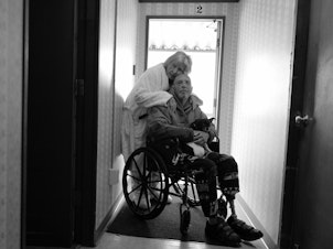 caption: Karen hugs Tony in his wheelchair on April 14, 2022. They now live in Burlington, Wash., and their mobile home no longer exists on the island. They are grateful to be together after being separated for long periods of time during the pandemic.