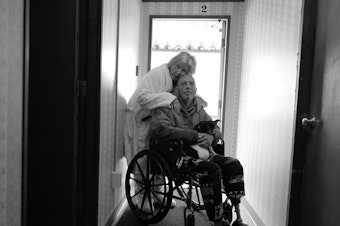 caption: Karen hugs Tony in his wheelchair on April 14, 2022. They now live in Burlington, Wash., and their mobile home no longer exists on the island. They are grateful to be together after being separated for long periods of time during the pandemic.