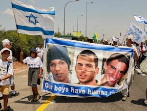 caption: Demonstrators marching with a banner showing the faces of captive Israeli civilian Avera Mengistu, and late soldiers Oron Shaul and Hadar Goldin from the Israeli city of Ashkelon arrive at the Karmia kibbutz on Aug. 5, 2022.