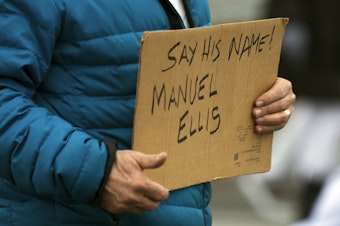 caption: 'Say His Name! Manuel Ellis,' reads a sign during a silent march of about 200 people honoring 33-year-old Manuel Ellis roughly one year after he was killed by Tacoma police officers on Sunday, February 28, 2021, along Martin Luther King Jr. Way in Tacoma. 