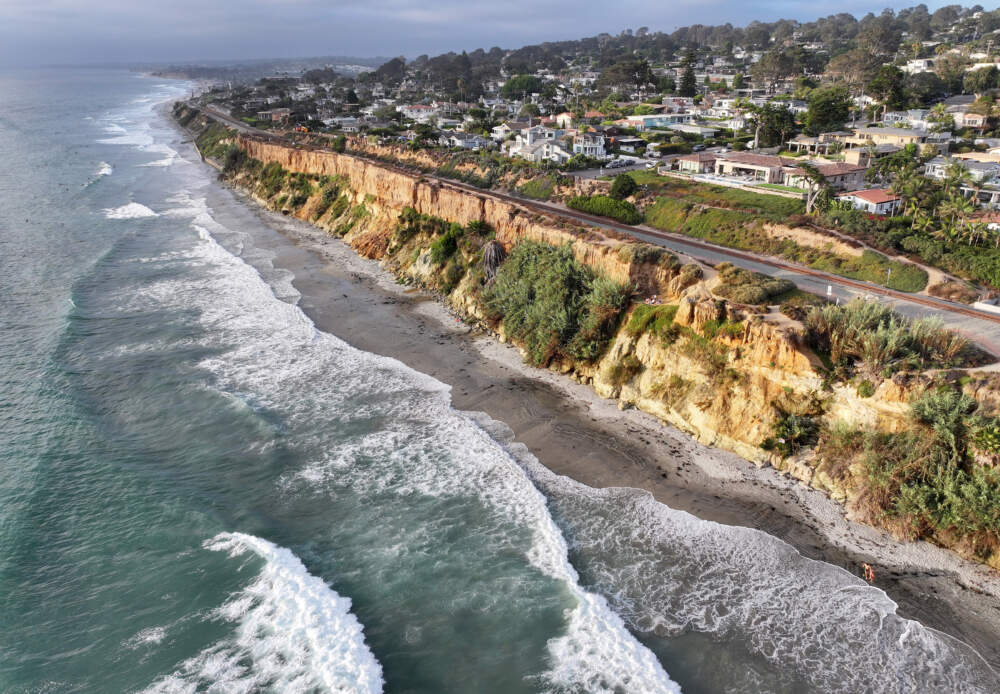 caption: An aerial view of waves breaking near eroding cliffs on the Amtrak Pacific Surfliner rail corridor at the Pacific Ocean coastline on August 16, 2024 in Del Mar, California. Rising sea levels and stronger storms have contributed to increased erosion and landslides along the vital Amtrak Pacific Surfliner coastal rail corridor which runs over 350 miles through Southern California to California’s Central Coast. Erosion and landslides on coastal bluffs led to three extended closures along the route in 2023 and 