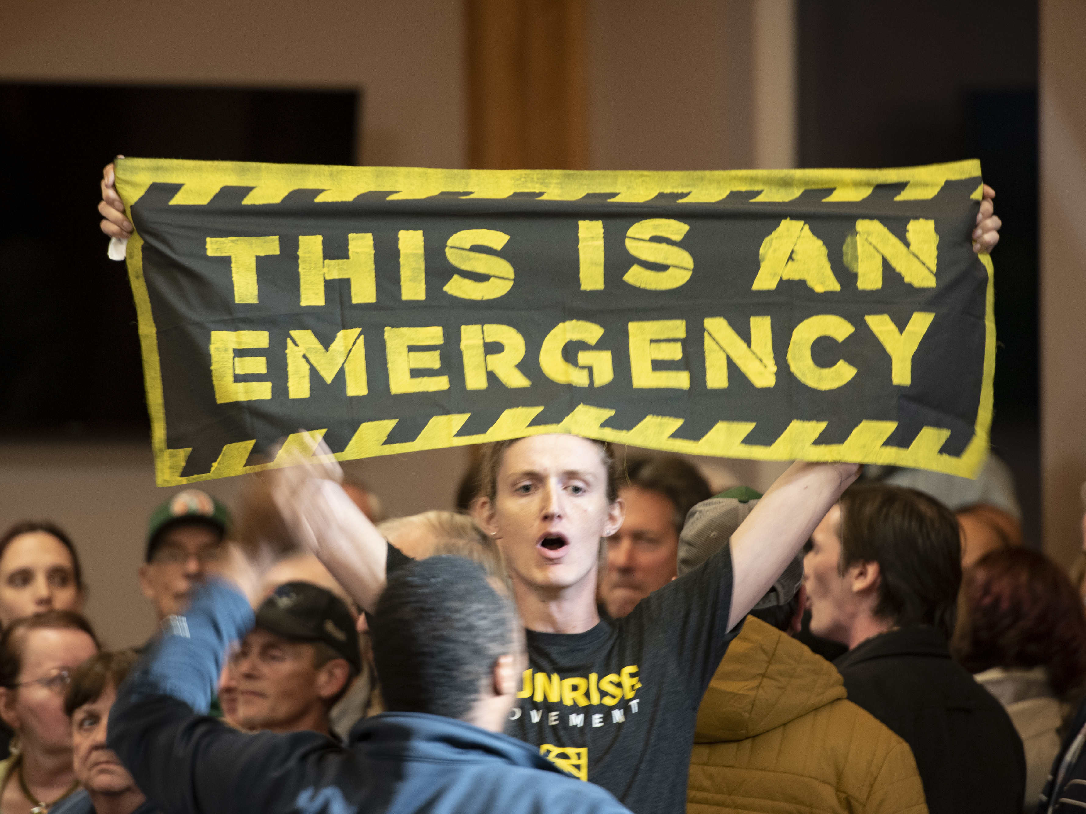 caption: A climate change protester holds a banner reading, "This Is An Emergency," during a 2019 town hall event in Manchester, N.H., with Joe Biden, who isn't pictured.