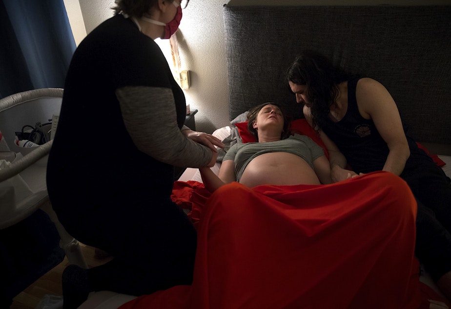 caption: Midwife Jenn Linstad, left, and Jake Black, right, hold Hope Black's hands as she breathes through a contraction at 2:25 a.m. on Friday, May 28, 2020, at the couple's home on Vashon Island.