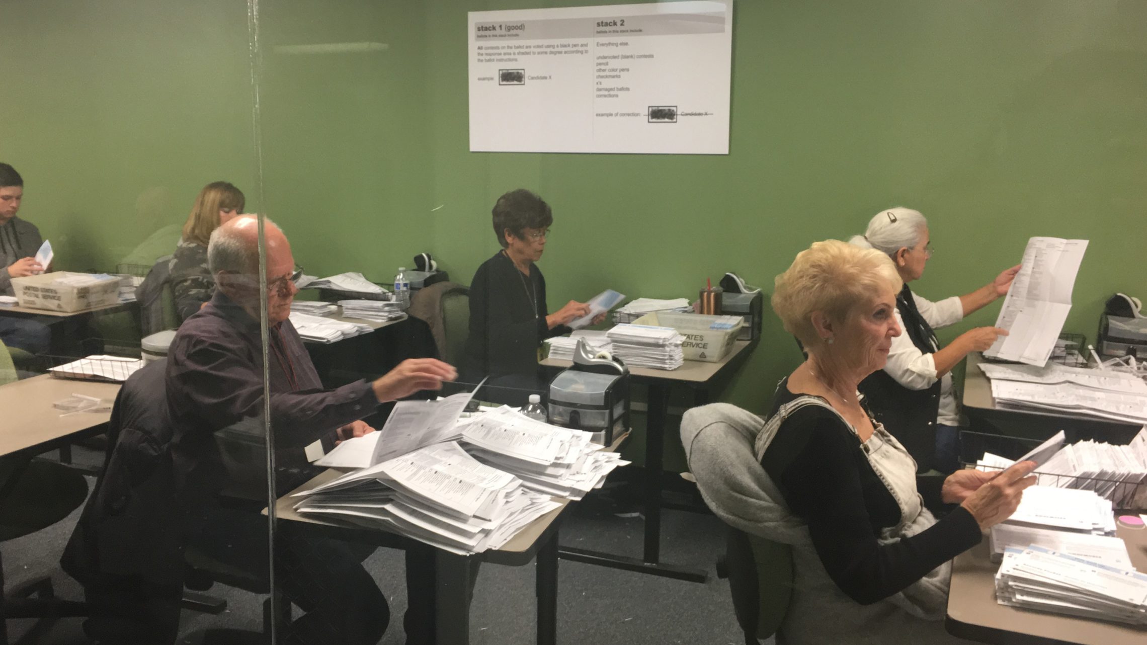caption: Workers in the Yakima County Elections Office work to open ballots and verify information. Their work is done within public view behind a glass wall, for anyone to monitor. CREDIT: ESMY JIMENEZ/NWPB