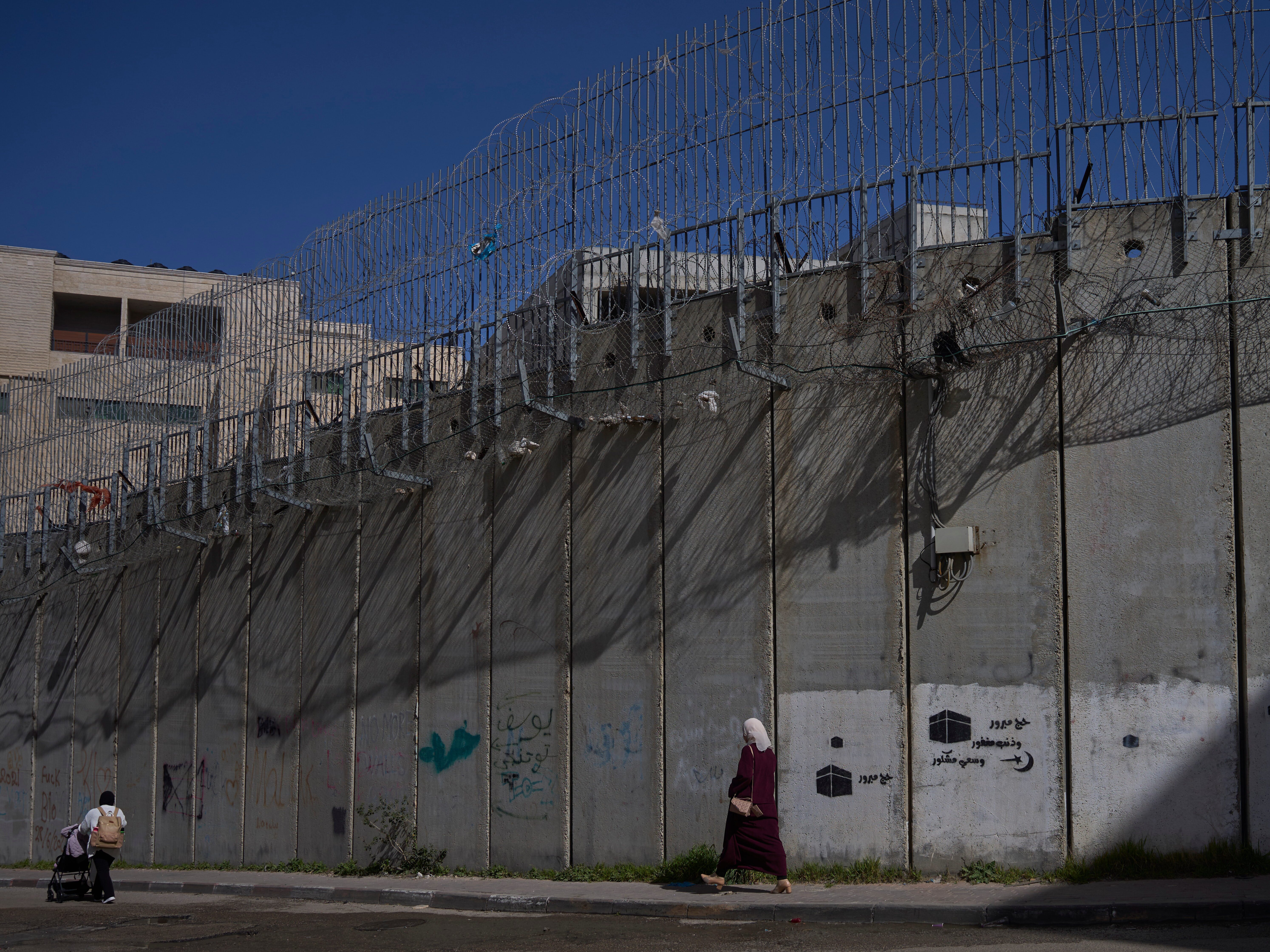 caption: Palestinians walk along the separation barrier between the West Bank and east Jerusalem neighborhood of Beit Hanina, Sunday Feb. 15, 2026.