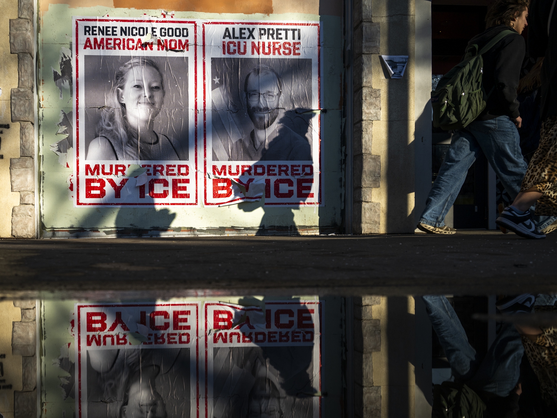 caption: People walk by signs showing the faces of Renee Good and Alex Pretti, the two U.S. citizens killed by federal immigration agents during the ICE surge in Minneapolis.