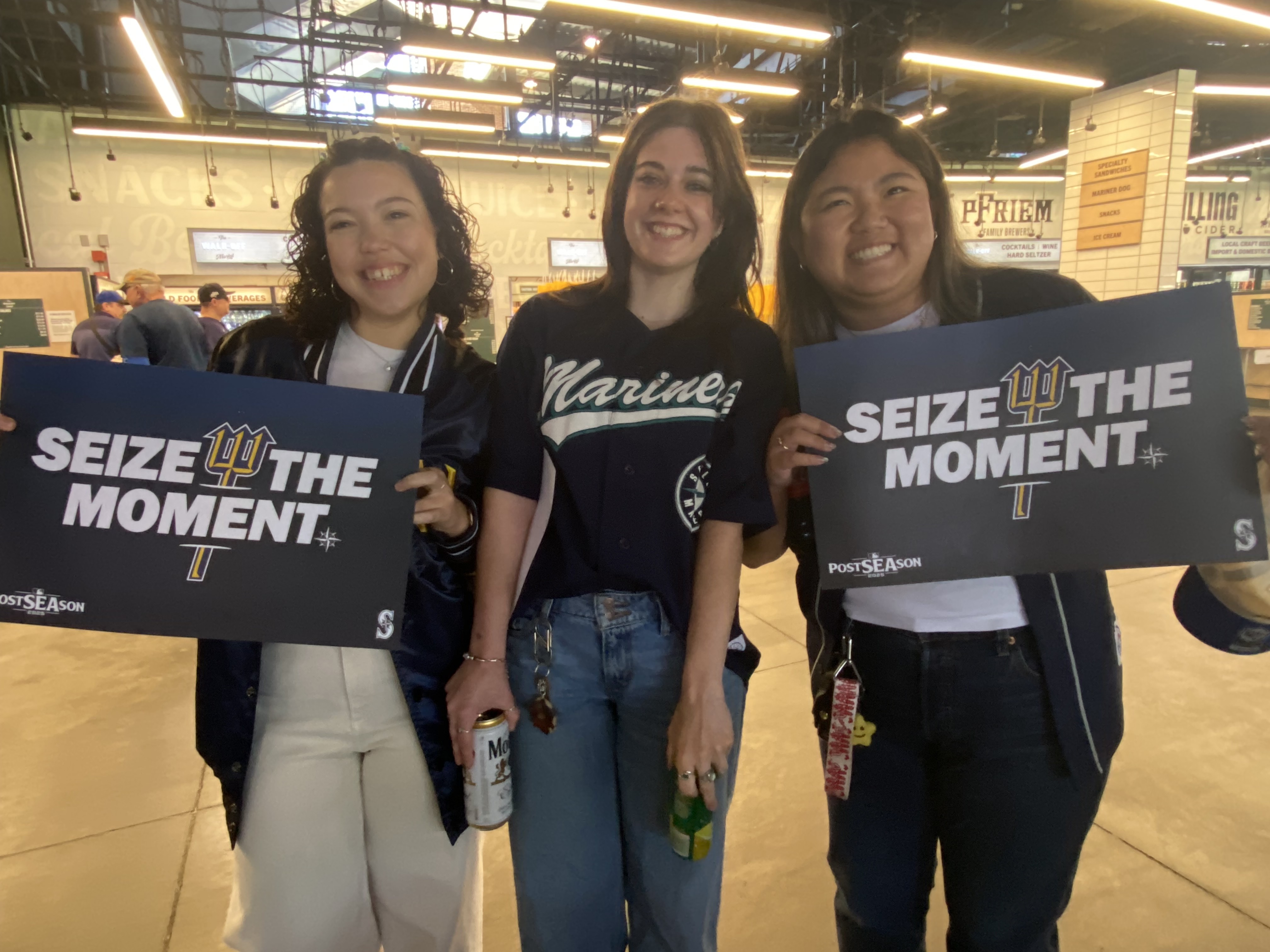 caption: Mariners fans Autumn Scheitlin (left), Zoe Ihrke (center), and Emily Hinrichsen (right) get ready to cheer on the team at a watch party held at T-Mobile Park.