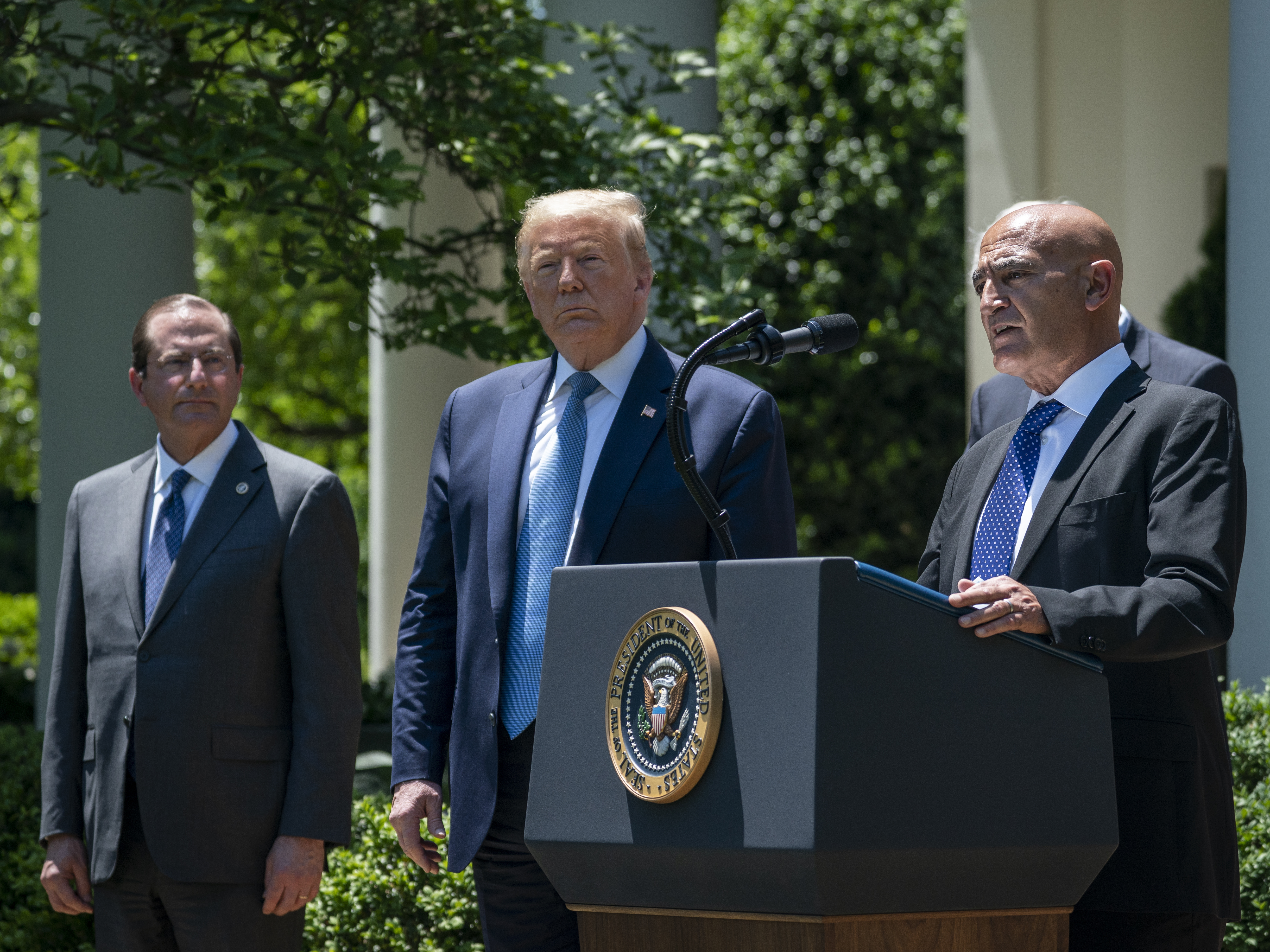 caption: Then-Health and Human Services Secretary Alex Azar (left) and President Donald Trump listen as Moncef Slaoui of Operation Warp Speed speaks about the crash program to develop a COVID-19 vaccine in the White House Rose Garden on May 15, 2020.