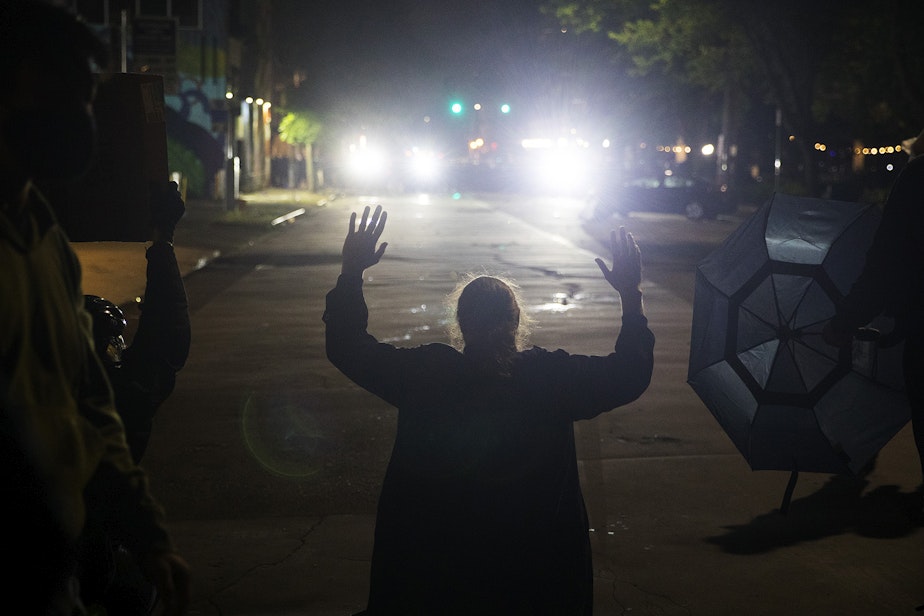 caption: A protester kneels with their hands in their air in front of hundreds of other protesters as Seattle police officers dressed in riot gear shine lights following the use tear gas, pepper spray, flash-bang grenades and rubber bullets on the fifth day of protests following the violent police killing of George Floyd on Tuesday, June 2, 2020, at the intersection 11th and Pine Streets in Seattle.