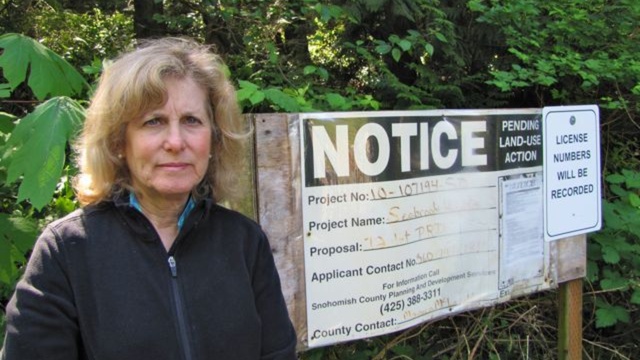 caption: Barbara Ingram stands in front of the site of a proposed development in her neighborhood. Snohomish County denied the permit for the development due to "slope stability and drainage issues" but the developer has reapplied.