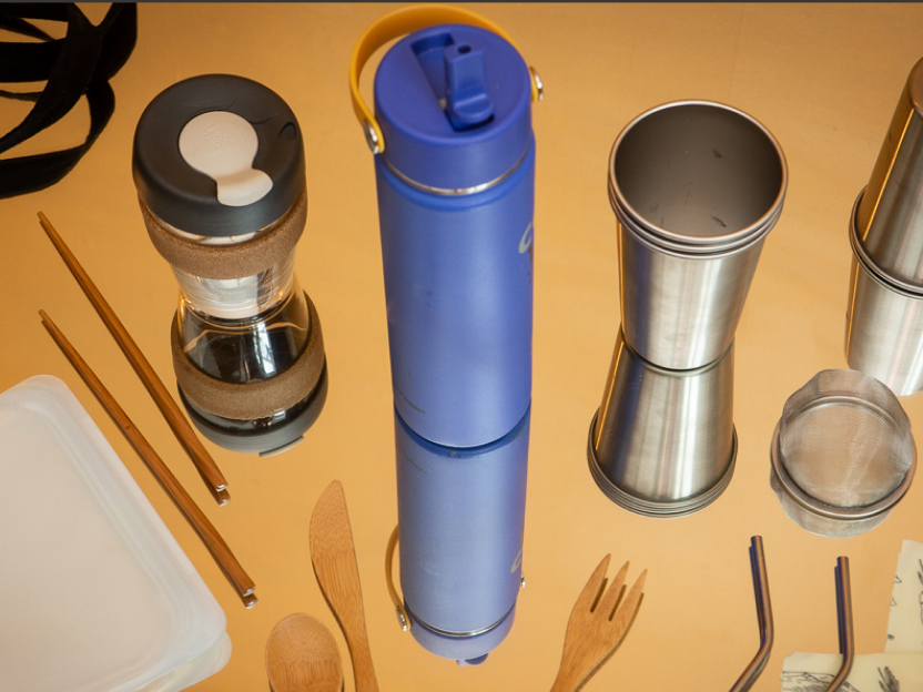 Photograph showing swaps for single-use plastics, including a metal water bottle, metal cup, bamboo flatware, glass coffee cup, silicone bag, metal straws, metal tea diffuser, bamboo chopsticks and canvas bag.