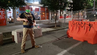 caption: A guard carrying an AR-15 rifle stands at the entrance to the CHAZ at 12th and Pike Street. 