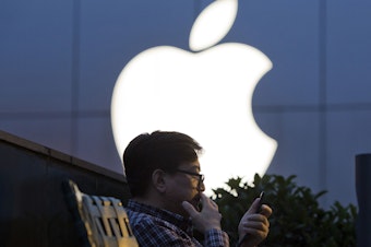 caption: A man uses his mobile phone near an Apple store logo in Beijing.