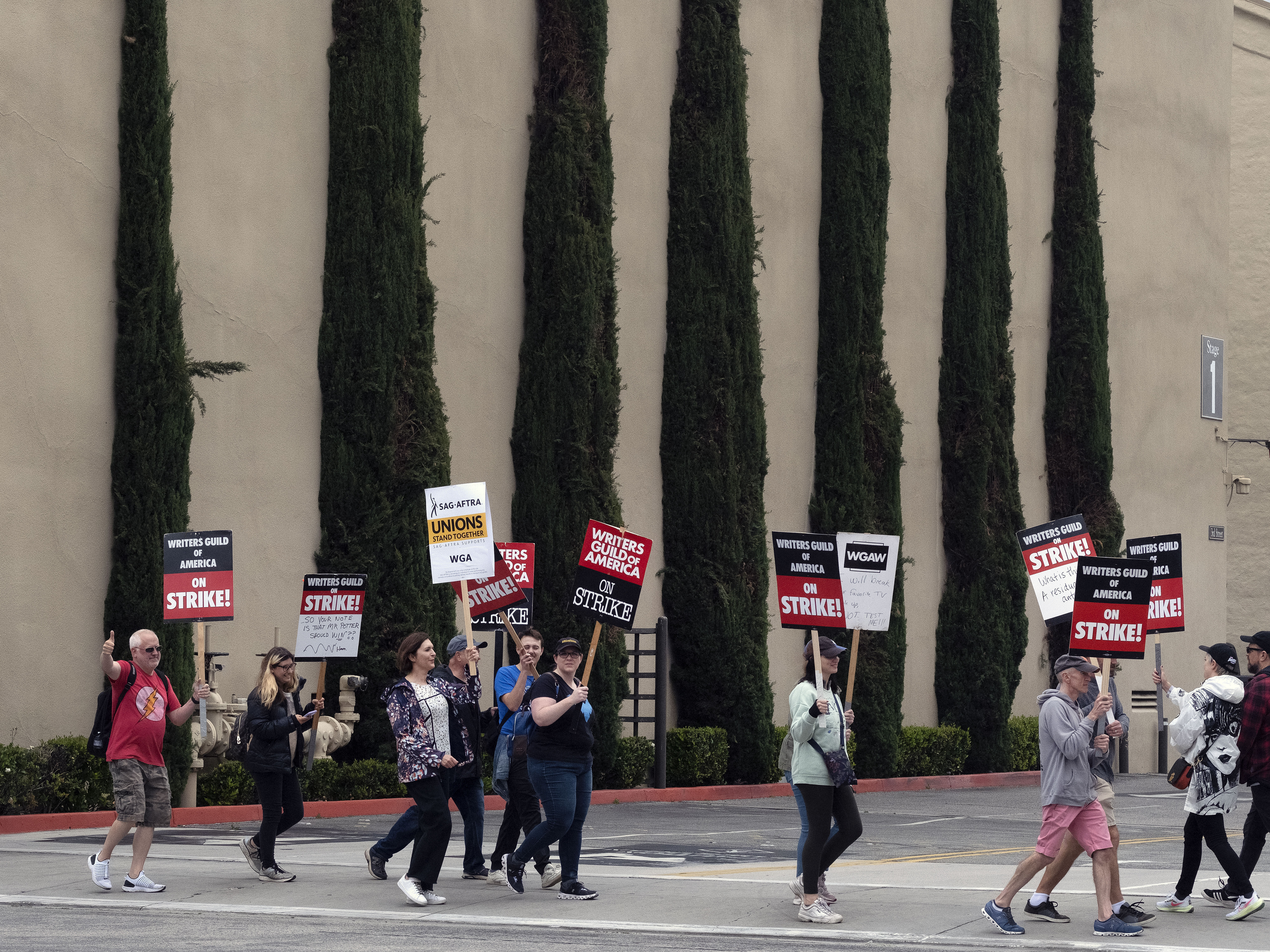 caption: Picketers pass near stage 5 at a studio entrance during a Writers Guild rally outside Warner Bros. Studios, on Wednesday, in Burbank, Calif.