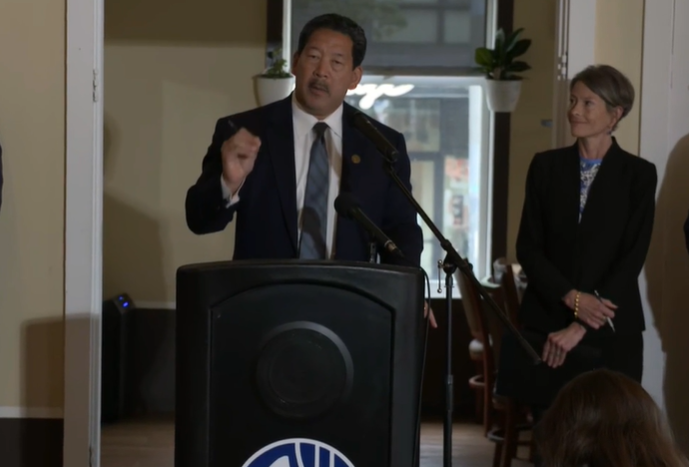 caption: Seattle Mayor Bruce Harrell stands next to Councilmember Sarah Nelson in the University District to introduce a new city program to help businesses recover after rash of broken windows and vandalism. 