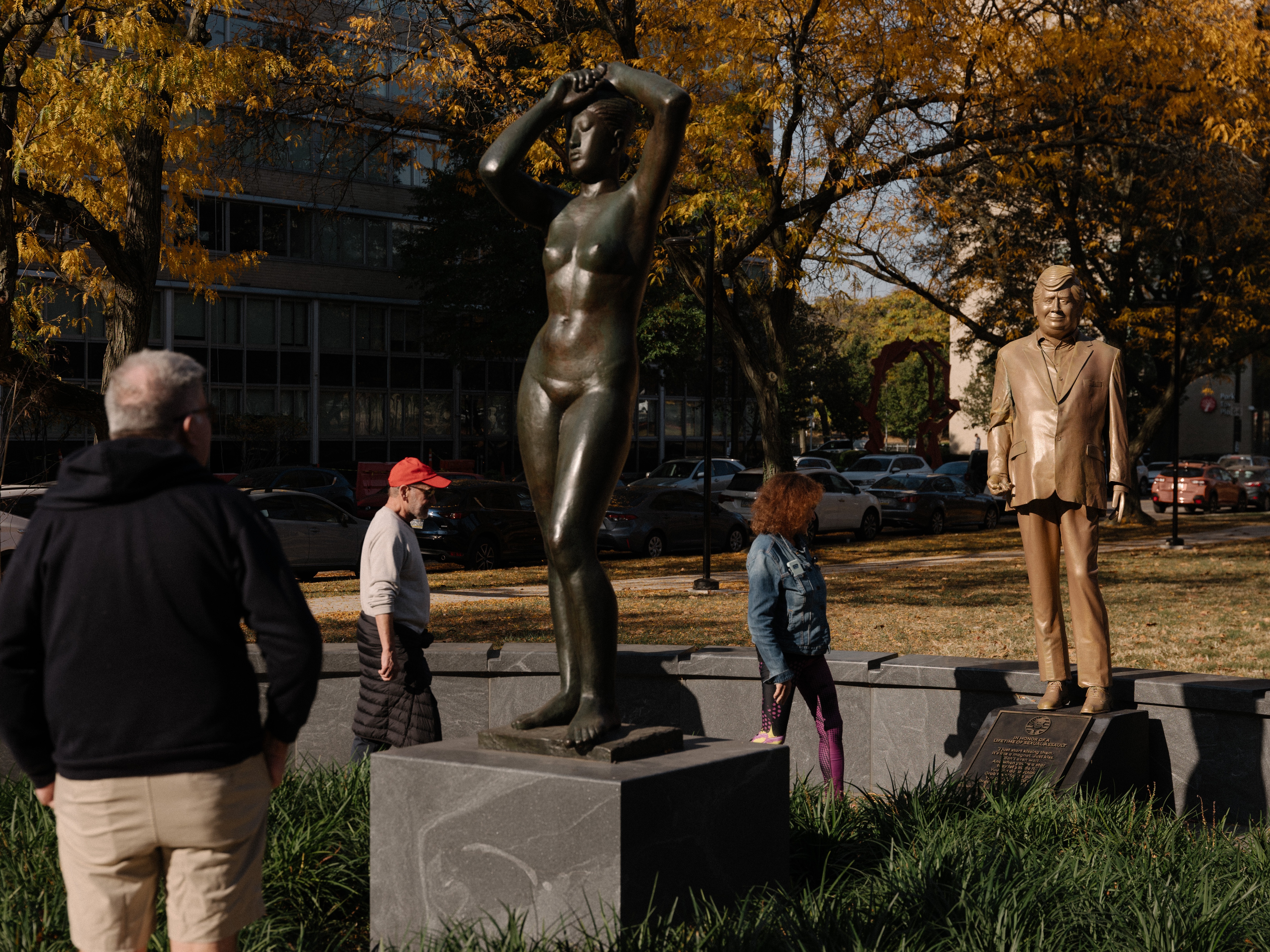 caption: Pedestrians look at a statue of Donald Trump behind Gerhard Marcks' sculpture <em>Maja</em>, in Maja Park in Philidelphia.