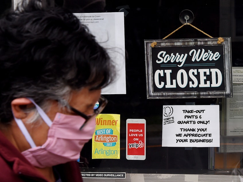 caption: A woman wearing a face mask walks past a sign in the window of a food store in Arlington, Va., Tuesday. Business groups say businesses are worried about liability issues as they reopen.